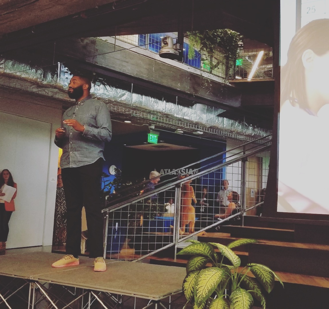 Jerold McDonald in a blue shirt standing on a small stage presenting to a room, with the venue's stairs and audience behind him